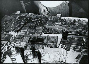 A black and white photo of a man with a lot of items on a table.